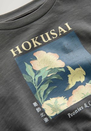 Hall kõrge kaelusega hall puuvillane dressipluus, millelt leiab Hokusai lillekunstiteose heledates roosades ja rohelistes toonides, sinise taustaga ja valge tekstiga, sealhulgas "Paeoniad ja Kameliaid."