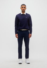 Άνδρας όρθιος, φοράει σκούρο μπλε πουλόβερ με άσπρο γιακά, ταιριαστό παντελόνι, λευκή ζώνη και λευκά αθλητικά παπούτσια, μπροστά σε απλό φωτεινό φόντο.