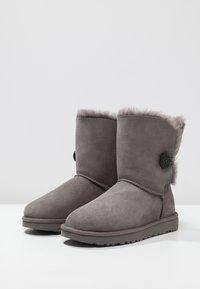 Stivaletti alla caviglia in suede grigio con punta rotonda, foderati in pelliccia sintetica, decorati con un bottone e con suola in gomma textured. Superficie liscia con finitura morbida.