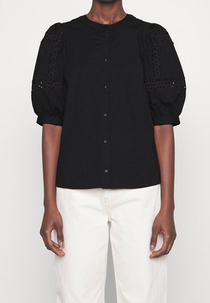 Person trägt eine schwarze, geknöpfte Bluse mit Spitzenärmeln und weißen Hosen, dargestellt vom Hals bis zur Hüfte vor einfachem Hintergrund.