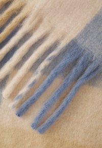 Nærbilde av myk ullstof med vekslende lysebeige og blå striper, med lange, fluffy frynser som gir tekstur.
