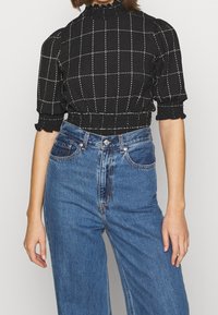Sort ternet cropped top med puffede ærmer og en rynket høj hals, parret med højtaljede blå denimjeans.