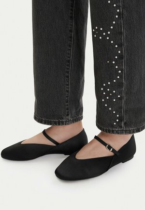 Zapatos planos negros con una delgada correa negra en el tobillo, usados con vaqueros gris oscuro que presentan pequeñas tachuelas plateadas en un patrón en zigzag en la parte baja de la pierna.