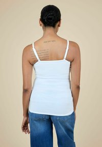 Balts, pieguļošs krekls bez piedurknēm ar plānām siksnām, prezentēts uz personas. Aizmugures skats rāda gludu tekstūru un tetovējumu pāri augšējai mugurai.