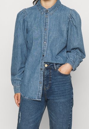 Kobieta ubrana w zapinaną na guziki niebieską długorękawową koszulę z bufkami na ramionach, w połączeniu z wysokimi niebieskimi dżinsami, z ręką w kieszeni.