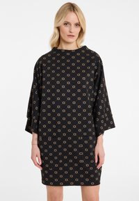 Zwarte jurk met een geometrisch gouden patroon, korte batwing mouwen en een relaxte pasvorm. Gemaakt van zachte stof met een gladde textuur.