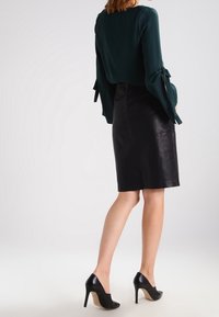 Femme debout, portant une blouse à manches longues vert foncé avec des poignets noués, une jupe en cuir noir jusqu'aux genoux et des chaussures à talons hauts noires.