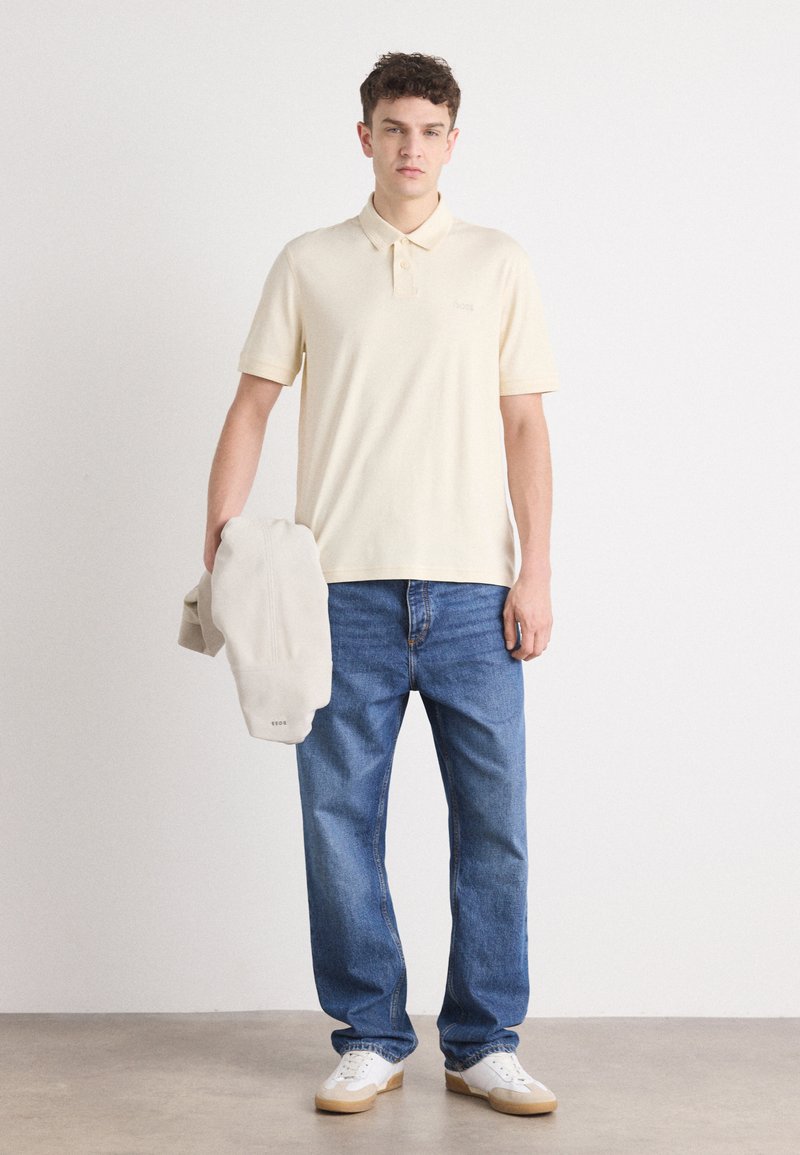 Polo shirt beige clair avec deux boutons, jeans bleus et baskets blanches avec des semelles beige. Tiens un sweat-shirt gris clair.