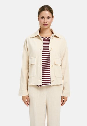 Beige Cordjacke mit einem Kragen, vorderem Knopfverschluss, zwei aufgesetzten Taschen und geripptem Stoff, getragen über einem gestreiften Hemd.