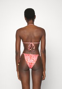 Conjunto de biquíni com tecido vermelho e um design estampado, cordas ajustáveis e acessórios em tom dourado. A vista traseira mostra uma cobertura mínima.