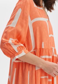Langärmliges orangenes Kleid mit weißen geometrischen Mustern, ausgestattet mit einer gerafften Taille und einem weichen, strukturierten Stoff. Knopfdetail am Ärmel.