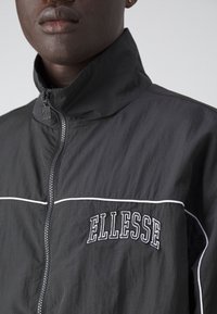Veste légère noire avec fermeture éclair, col montant, lignes d'accent blanches, et logo "ELLESSE" brodé sur la poitrine. Texture lisse.