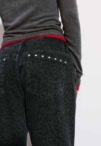 Črne denim hlače z diskretno leopardjim vzorcem, srebrni nitasti dodatki na žepih in rdeč pas, ki se pokaže pod sivim puloverjem.