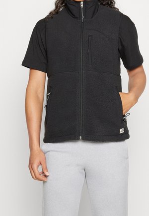 Person trägt eine schwarze Fleeceweste mit Reißverschluss über einem schwarzen Kurzarmshirt und hellgraue Jogginghosen, Hände locker seitlich.