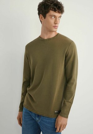 Młody mężczyzna patrzący w prawo, ubrany w oliwkowozieloną koszulę z długim rękawem i niebieskie dżinsy na jasnym, jednolitym tle.