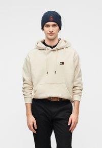 Μπεζ βαμβακερό φούτερ με κουκούλα, εμπρός τσέπη και μικρό λογότυπο. Συνδυάζεται με μαύρο παντελόνι και μπλε πλεκτό σκουφάκι με κόκκινο σήμα.