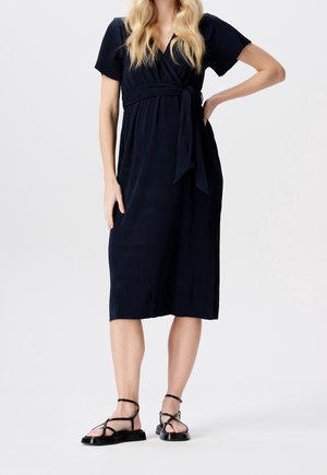 Nainen, joka pukeutunut mustaan, polvipituiseen, lyhythihaiseen kietaisumekkoon, jossa on vyötärönauha, ja mustiin remmikenkäisiin tasapohjaisiin sandaaleihin, seisoo valkoista taustaa vasten.