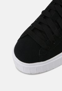 Gros plan sur le bout noir d'une basket avec des lacets noirs sur une épaisse semelle en caoutchouc blanche sur un fond gris.