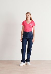Camiseta de cuello en V rosa con logo, jeans de mezclilla oscuros de cintura alta y zapatillas blancas. Tela suave, ajuste estructurado y diseño clásico.