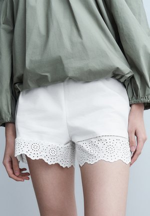 Zielona bluzka z długim rękawem w zestawieniu z białymi szortami ozdobionymi misterną koronką typu eyelet na krawędzi, noszona przez osobę stojącą z luźno opuszczonymi ramionami.