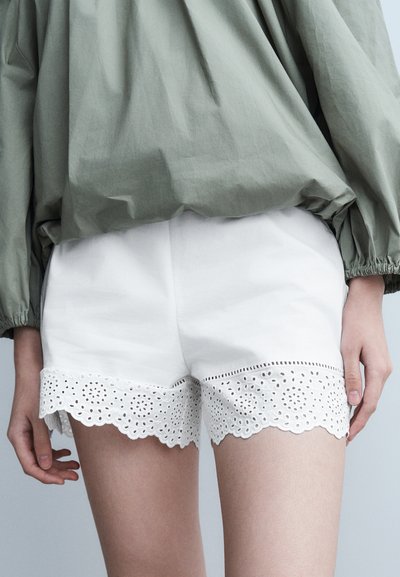 Haut vert à manches longues associé à un short blanc orné d'une bordure en dentelle ajourée intricate à l'ourlet, porté par une personne debout, les bras détendus.