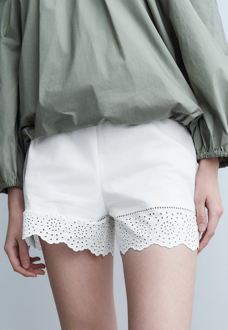 Πράσινο μακρυμάνικο τοπ σε συνδυασμό με λευκά σορτς διακοσμημένα με περίτεχνη δαντέλα αυγότρυπα στο κάτω μέρος, φορεμένα από ένα άτομο που στέκεται με χαλαρά τα χέρια.