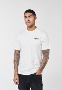 Mand med kort hår og tatoveringer på begge arme iført hvid t-shirt med grafik og sorte bukser, stående mod en ensfarvet baggrund.