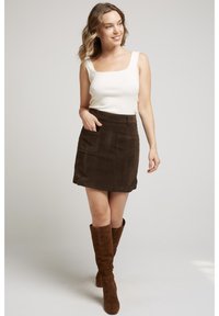 Jeune femme souriante, portant un haut sans manches blanc, une jupe marron foncé avec des poches et des bottes montantes assorties, se tenant sur un fond uni.