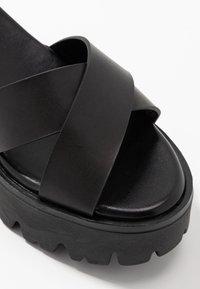 Sandale à plateforme noire avec des lanières en simili cuir croisées. Présente une semelle épaisse et texturée ainsi qu'un footbed lisse et mat.