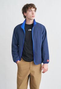 Ναυτικό μπουφάν με μπλε λεπτομέρειες, κλείσιμο με φερμουάρ και λεπτομέρειες από φλις στο γιακά. Φορεμένο πάνω από μαύρο t-shirt και συνδυασμένο με ανοιχτά καφέ παντελόνια.