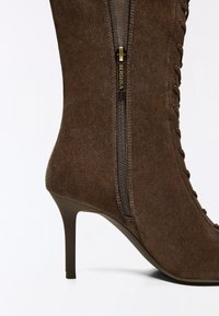 Botte à talon haut en daim marron avec fermeture éclair latérale et détails à lacets sur le côté, dotée d'un talon stiletto fin.