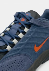 Tenisky Nike s modrou a šedou síťovanou horní částí, oranžovým logem Swoosh, texturovanou tkaninou a bílými tkaničkami s kontrastními akcenty.