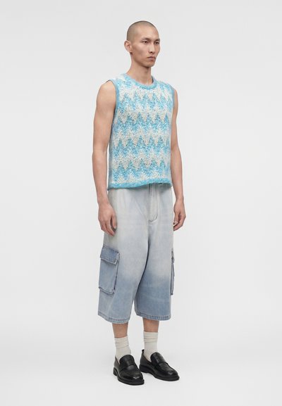 Άνδρας στέκεται φορώντας αμάνικο μπλε και λευκό πλεκτό τοπ με ζικ-ζακ σχέδιο, χαλαρά ανοιχτόχρωμα τζιν cargo σορτς, λευκές κάλτσες και μαύρα μοκασίνια σε λευκό φόντο.