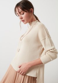 Beige kootud sviiter kõrge kaelusega ja külgmiste lõigetega, koos pleeditud beežist seelikuga. Aksessuaariks on kihiline kett. 