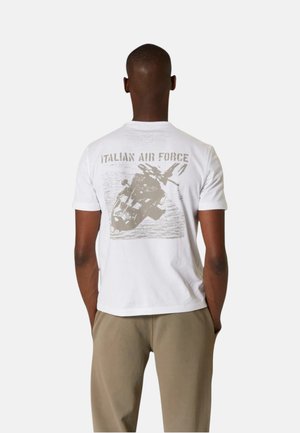 Uomo che indossa una t-shirt bianca con il testo "Italian Air Force" e una grafica di un elicottero, abbinata a pantaloni beige.