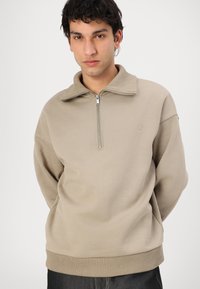 Sudadera beige de media cremallera con un alto cuello y puños acanalados. Fabricada en un suave tejido texturizado, presenta un pequeño logo en el pecho.