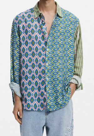 Mann trägt ein langärmliges Button-up-Hemd mit gemischten blauen, grünen und pinkfarbenen geometrischen und gestreiften Mustern sowie eine hellblaue Jeans mit Stickerei.