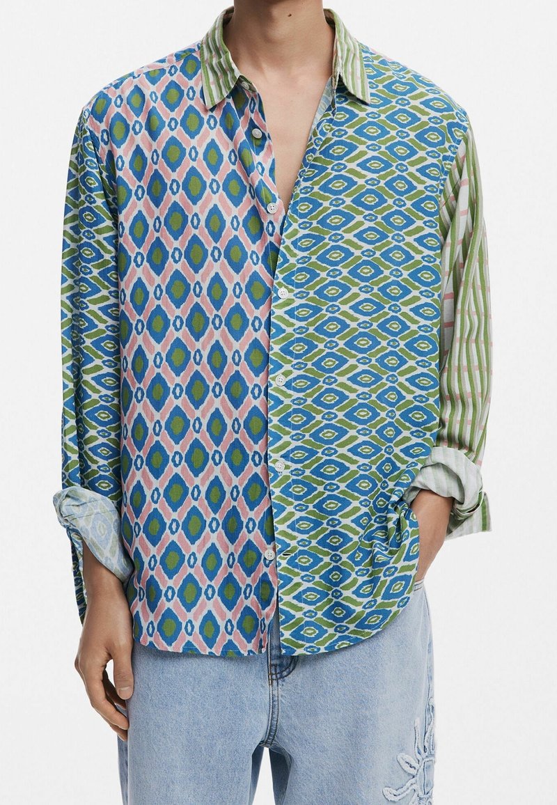 Homme portant une chemise à manches longues boutonnée, aux motifs géométriques et rayés mêlant bleu, vert, rose, et un jean bleu clair brodé.