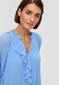 Lichtblauwe blouse met geplooide textuur, een gerimpelde kraag en detail aan de voorkant, met lange mouwen en een V-halslijn. Zacht, lichtgewicht stof.