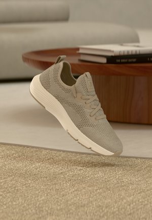 Beżowy dziergany sneaker z białą podeszwą, sznurowadłami i pętelką, unoszący się nad teksturowanym dywanem w pobliżu drewnianego stolika kawowego z poukładanymi książkami.