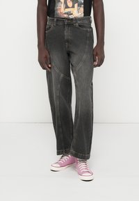 Άτομο που φοράει μαύρα ξεφτισμένα παντελόνια σε ευρεία γραμμή, ένα γραφικό μαύρο T-shirt και μωβ καμβά sneakers με ψηλή μπότα, στέκεται σε λευκό δάπεδο.