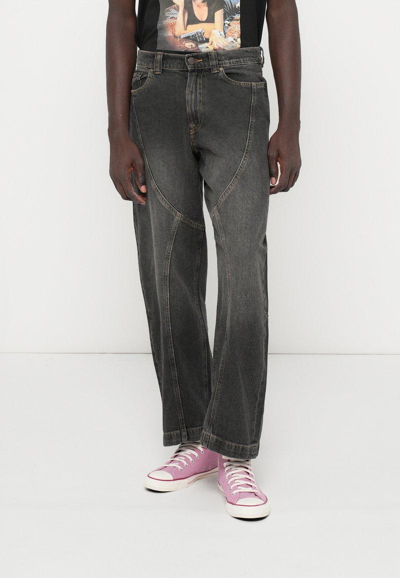 Άτομο που φοράει μαύρα ξεφτισμένα παντελόνια σε ευρεία γραμμή, ένα γραφικό μαύρο T-shirt και μωβ καμβά sneakers με ψηλή μπότα, στέκεται σε λευκό δάπεδο.