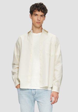 Crème-kleurig shirt met knoopsluiting aan de voorkant, lange mouwen en een borstzak, gelaagd over een plain wit T-shirt. Draagt het met lichtblauwe jeans.