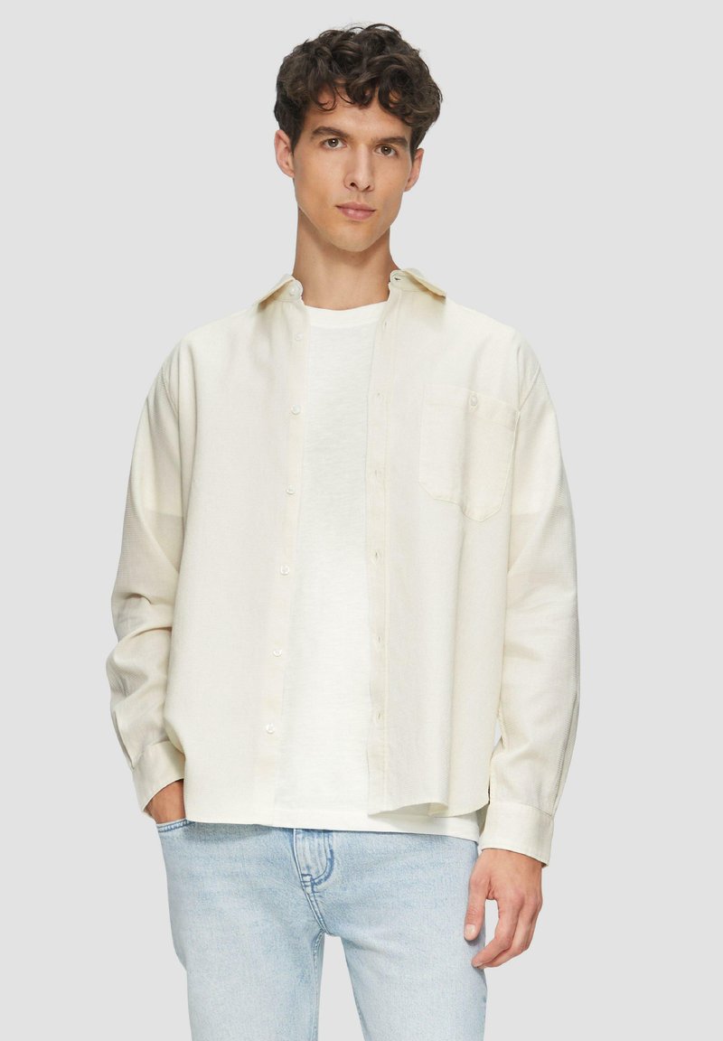 Crème-kleurig shirt met knoopsluiting aan de voorkant, lange mouwen en een borstzak, gelaagd over een plain wit T-shirt. Draagt het met lichtblauwe jeans.