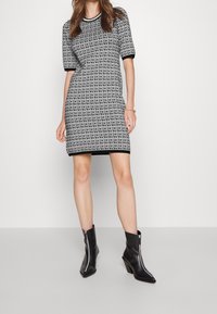 Femme portant une robe à manches courtes noire et blanche imprimée du logo DKNY, associée à des bottines noires à bouts pointus avec des fermetures éclair à l'avant.