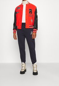 Man i en röd universitetsjacka med svarta ärmar, vit polo-tröja, marinblå joggers och beiga ankelstövlar som står mot en vit bakgrund.