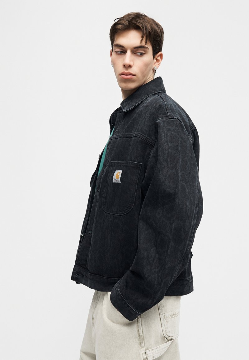 Jonge man die een zwarte jas met patroon draagt met een logo op de voorkant en lichtbeige broek, kijkt opzij tegen een effen achtergrond.