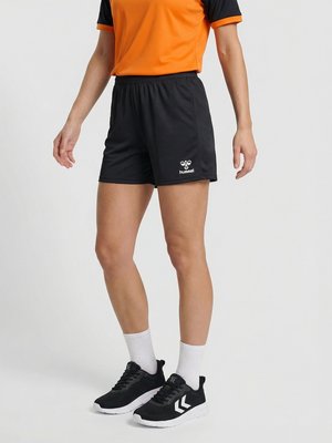 Person trägt schwarze Sportshorts mit Hummel-Logo, orange-schwarzes kurzärmeliges Shirt, weiße Socken und schwarze Sportschuhe und steht vor weißem Hintergrund.