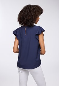 Blusa blu navy con maniche arricciate, dettaglio a chiave circolare nella parte posteriore, tessuto liscio e orlo curvo, abbinata a pantaloni bianchi.
