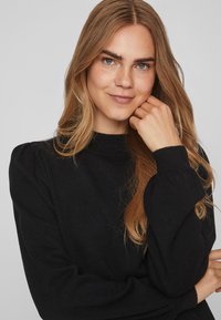 Schwarzer Strickpullover mit hohem Kragen, langen Ärmeln und leicht gepolsterten Schultern. Die Textur wirkt weich und glatt.